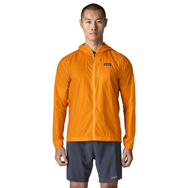 En person står med ansigtet vendt fremad foran en ensfarvet hvid baggrund og har en Patagonia Houdini-jakke (herre) i farven Autumn Orange på samt grå shorts.