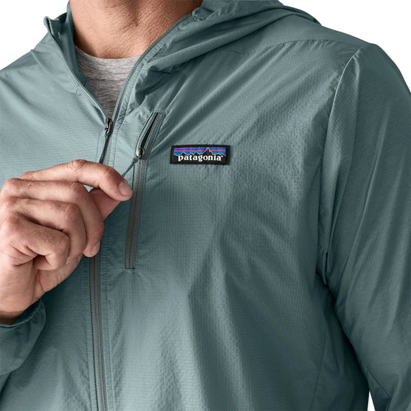 En person er iført en Patagonia Houdini Jacket, Herre, i Blue Sage, fremstillet af genanvendt nylon. Hånden trækker lynlåsen op, og Patagonia-logoet er synligt på brystet.
