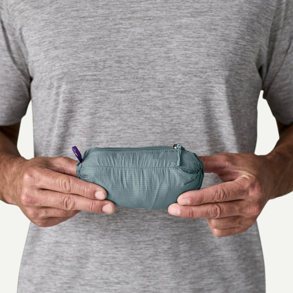 En person i grå t-shirt holder en lille blågrøn pose lavet af genbrugsnylon. Ligesom Patagonias Houdini Jacket, Herre, Blue Sage, har den en lilla løkke og er let og praktisk.