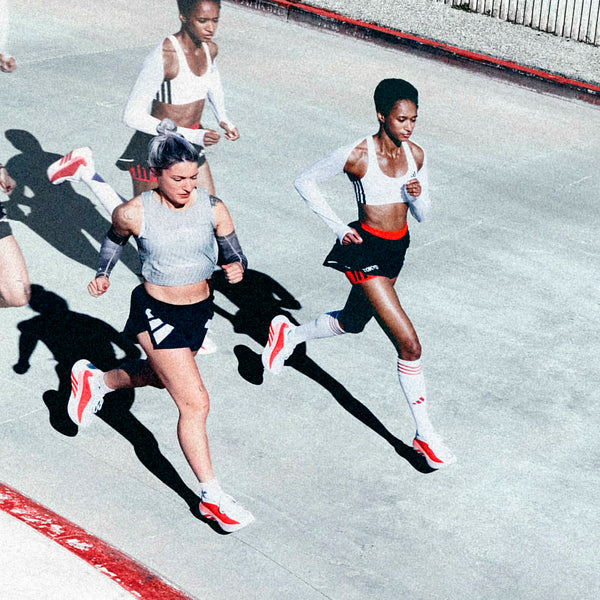 Fire kvinder i sportstøj, alle iført Adidas Hyperboost Edge Dame i Cloud White/Pure Ruby/Zero Metalic, løber på en udendørs bane. Fotograferet ovenfra strækker deres lange skygger sig over fortovet ved siden af et hegn.