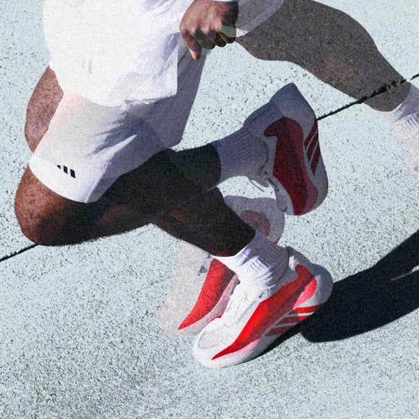 En løber i hvide shorts, sokker og Adidas Hyperboost Edge, Herre (Cloud White/Pure Ruby/Zero Metalic) går på struktureret beton; en stilfuld scene, der fremhæver komforten og ydeevnen i disse Adidas landevejsløbesko.