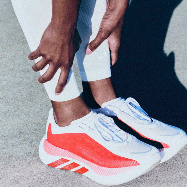 En kvinde i hvid Adidas Hyperboost Edge (Cloud White/Pure Ruby/Zero Metalic) justerer manchetten på sine hvide bukser, mens hun står på beton, klar til behageligt løb på vejen.