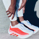 En kvinde i hvid Adidas Hyperboost Edge (Cloud White/Pure Ruby/Zero Metalic) justerer manchetten på sine hvide bukser, mens hun står på beton, klar til behageligt løb på vejen.