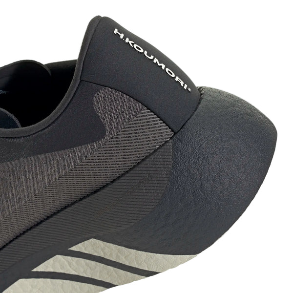 Nærbillede af hælen og siden på en sort Adidas Hyperboost Edge-sneaker, Dame, Chacoa/Ntgrey/Alumin x Hermanos Koumori, med påskriften »H. KOUMORI« på fligen bagpå. Denne sko minder om de komfortfokuserede løbesko til landevejsløb, som man finder i Hyperboost Edge-kollektionen, og har en struktureret sål samt et diskret stribet design langs siden.