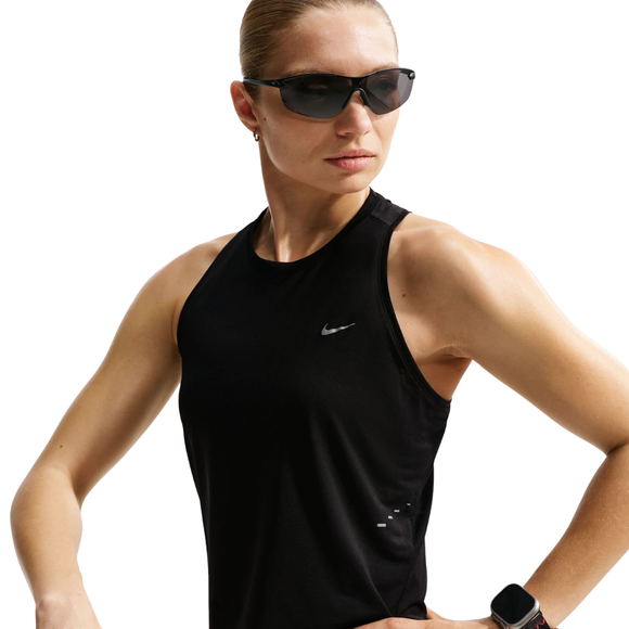 En kvinde i mørke solbriller og en sort Nike Nk Swift Nvlt Brthe Tank (Black/White/Reflective Silver) står med hænderne på hofterne og kigger til siden. Hun har et smartwatch på venstre håndled. Den ensfarvede baggrund er hvid.