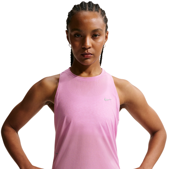 En kvinde med flettet hår bærer en ærmeløs Nike Nk Swift Nvlt Brthe Tank til kvinder i Lt Magenta/Photon Dust/Reflective Silv, lavet af åndbart stof, og står med hænderne på hofterne mod en hvid baggrund.