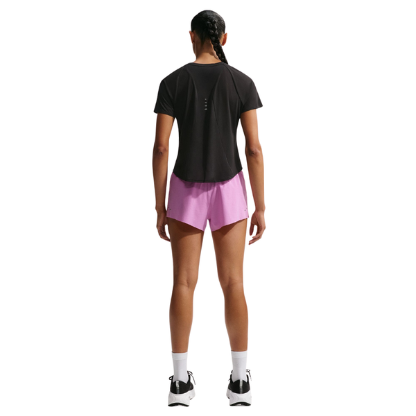 En person med flettet hestehale, klædt i sort t-shirt, Nike Swift Breathe Mid-Rise 2.5" løbeshorts til kvinder i Lt Magenta/Photon Dust/Reflective Silver, hvide sokker og sort-hvide sneakers står med ansigtet vendt væk.