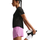 En kvinde i sort T-shirt, Swift Breathe Mid-Rise 2.5" Running Shorts (Lt Magenta/Photon Dust) fra Nike, hvide sokker og sort-hvide sportssko strækker sit venstre ben ud på en almindelig hvid baggrund. Shortsene har Dri-FIT-teknologi.