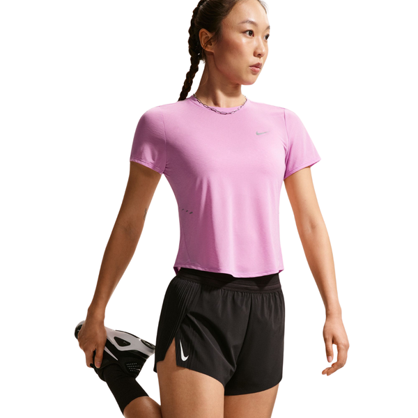 En kvinde i en pink Nk Swft Nvlt Brthe Ss Top, Dame by Nike og sorte shorts strækker benet og holder sig kølig i denne Dri-FIT løbe-T-shirt mod en almindelig hvid baggrund.