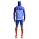 En person ser væk og er iført en blå jakke med hætte, Nike Stride Running Half Tights Herre i Lapis/Reflective Silver, orange sokker og orange sportssko på en almindelig hvid baggrund.