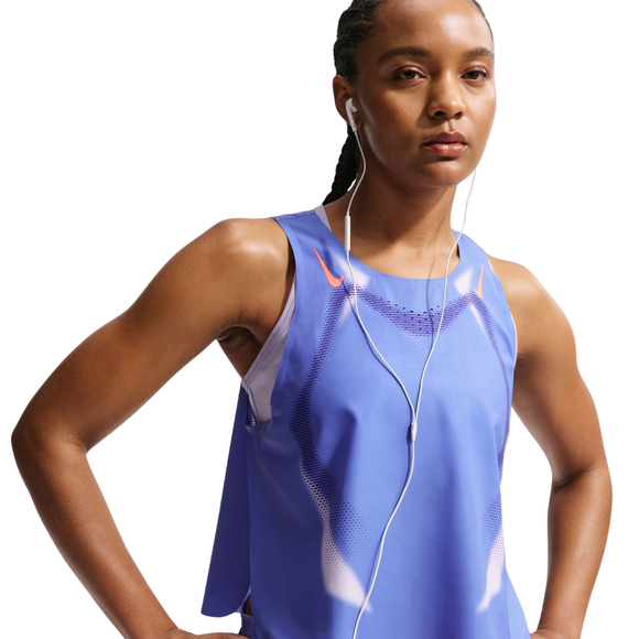En kvinde i Nike Nk Arswft Dfadv Crop Tank Gx, Dame, Sapphire/Hot Lava/Hot Lava med race-fit crop-design og hvide høretelefoner står med hænderne på hofterne og ser frem for sig. En almindelig hvid baggrund fremhæver Dri-FIT ADV-teknologien.