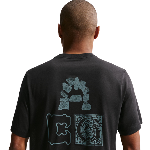 En person med glatbarberet hoved ses bagfra iført en sort Nike ACG Dri-FIT T-shirt til mænd, som har tre blå grafiske mønstre på ryggen, herunder stenformer, et rektangel og et cirkulært mønster.