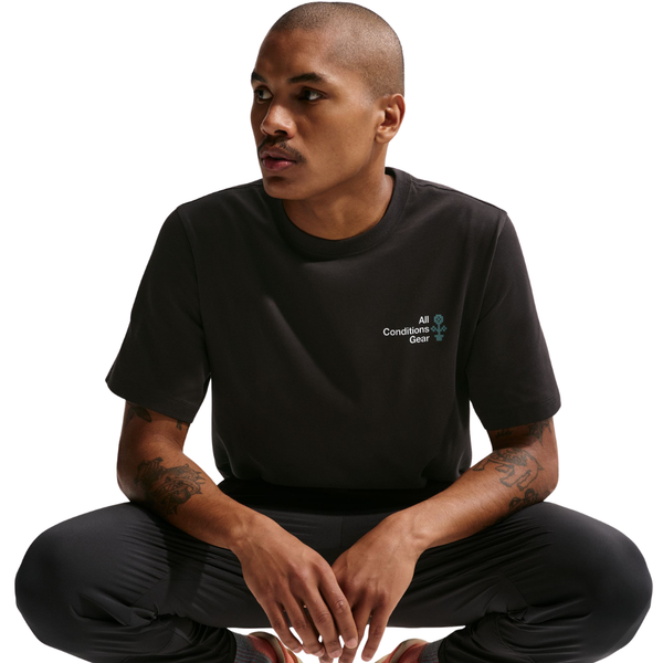 En tatoveret, korthåret mand sidder på hug i sorte bukser og en Nike ACG Dri-FIT T-shirt, Herre, Black, der poserer mod en almindelig hvid baggrund.