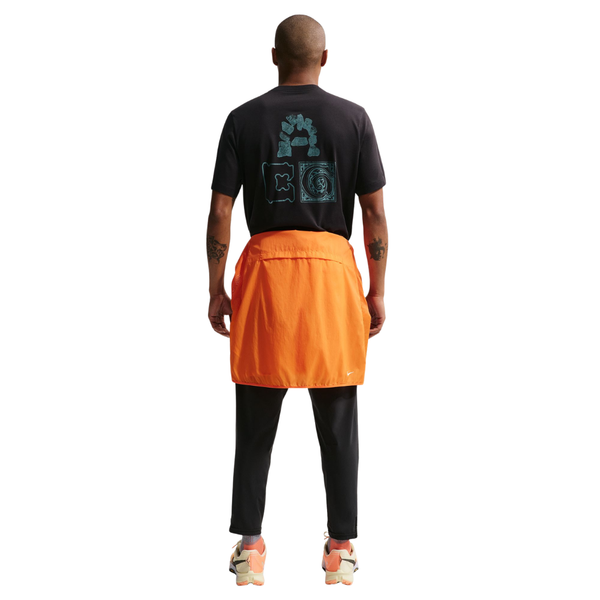 En person kigger væk iført sort Nike ACG Dri-FIT T-shirt (Herre), sorte bukser, en orange jakke bundet i taljen og lyse sneakers med orange detaljer på en hvid baggrund.