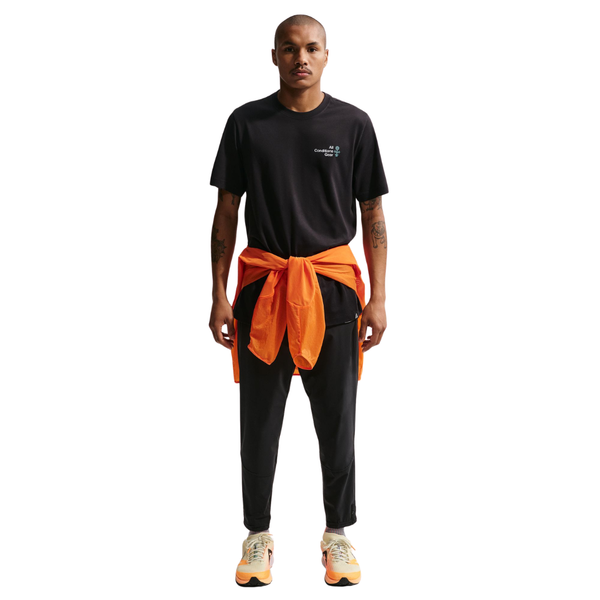 En person kigger fremad i en sort Nike ACG Dri-FIT T-shirt, Herre, sammen med sorte bukser og lyse sneakers med orange detaljer. En orange jakke er bundet i taljen, alt sammen på en almindelig hvid baggrund.