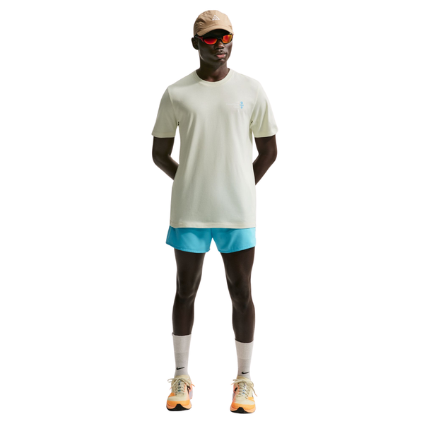 En person står og kigger fremad iført beige kasket, orange solbriller, Nike ACG Dri-FIT T-Shirt Herre Sea Glass, lyseblå shorts, beige sokker og beige og orange løbesko på en hvid baggrund.
