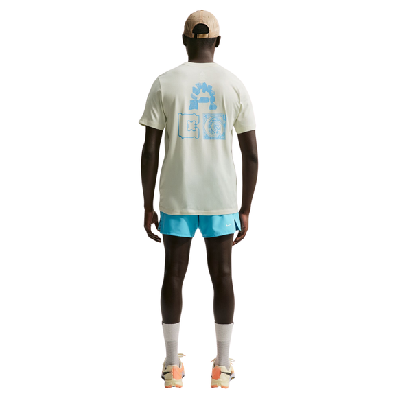 En person kigger væk, iført en beige kasket, Nike ACG Dri-FIT T-Shirt Herre Sea Glass med blå ryggrafik, blå shorts, hvide sokker og beige sneakers med orange detaljer - et ideelt udendørs look til mænd på en hvid baggrund.