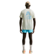 En person kigger væk, iført en beige kasket, Nike ACG Dri-FIT T-Shirt Herre Sea Glass med blå ryggrafik, blå shorts, hvide sokker og beige sneakers med orange detaljer - et ideelt udendørs look til mænd på en hvid baggrund.