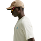 En person iført en beige baseballkasket, røde solbriller og en Nike ACG Dri-FIT T-Shirt Herre in Sea Glass med blåt logo på brystet står i profil mod en ensfarvet baggrund.