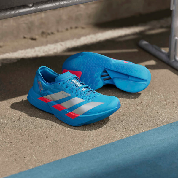 Et par Adidas Adizero Adios Pro 4 herre løbesko i Solblu/Silvmt/Lucred er placeret på beton ved siden af et blåt spor - det ene opretstående, det andet på siden, så man kan se sålen.