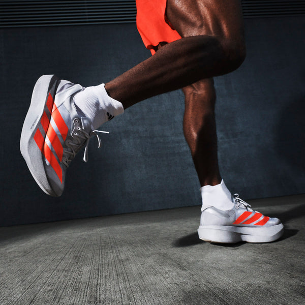 Nærbillede af en persons ben i Adidas Pro Evo 2 Unisex-sko (Silver Metallic/Lucid Red/Grey Two), hvide sokker og orange shorts med den ene fod løftet fra jorden på en mørk baggrund af beton.