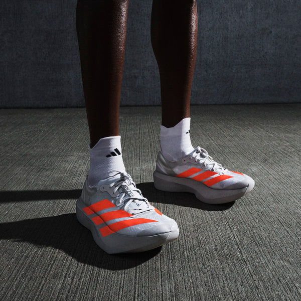 En person iført Adidas Pro Evo 2 unisex-sko i sølvmetallic/lucidrød/grå to står på en struktureret grå overflade med en mørk betonbaggrund, parret med hvide Adidas-sokker.