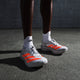 En person iført Adidas Pro Evo 2 unisex-sko i sølvmetallic/lucidrød/grå to står på en struktureret grå overflade med en mørk betonbaggrund, parret med hvide Adidas-sokker.