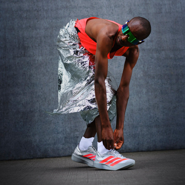 Iført en sølvmetallic nederdel, rød top, grønne solbriller med visir og Adidas Pro Evo 2 sko (Unisex, Silver Metallic/Lucid Red/Grey Two) bøjer en person sig ned for at binde sin sko mod en grå tekstureret vægbaggrund.