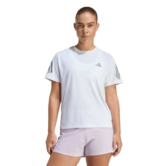 En kvinde med tilbagestrøget hår og et neutralt udtryk ser frem for sig i en almindelig hvid omgivelse iført Adidas Adi365 Running Tee, Dame, White og lyserøde shorts - perfekt til løbetræning. Trøjen har et lille Adidas-logo.