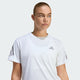 En kvinde med lys hud og tilbagestrøget blondt hår er iført Adidas Adi365 Running Tee, Dame, White - ideel til løbetræning - med grå logoer og står på en almindelig lysegrå baggrund.