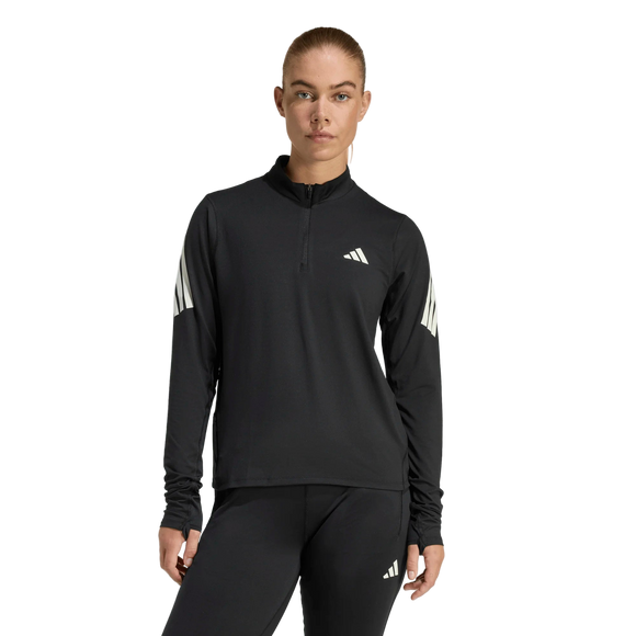 En person står med ansigtet fremad i Adidas Adi365 Zip Long Sleeve Shirt, Dame, Black. Den sorte top har en krave med kvart lynlås, hvide striber på begge ærmer og et hvidt Adidas-logo på venstre bryst. Matchende sorte leggings fuldender looket.