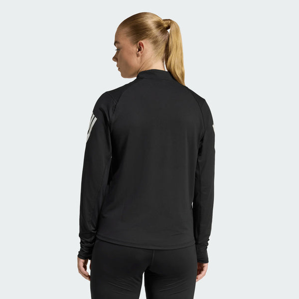 En kvinde med blondt hår i hestehale ses bagfra iført Adidas Adi365 Zip Long Sleeve Shirt til kvinder i sort. Hun sætter den sammen med sorte leggings på en almindelig, lys baggrund.