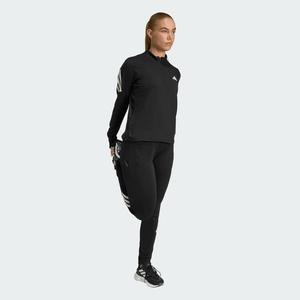 Iført Adidas Adi365 Zip Long Sleeve Shirt i sort står en person på ét ben, strækker sit baglår og kigger fremad mod en almindelig lys baggrund.