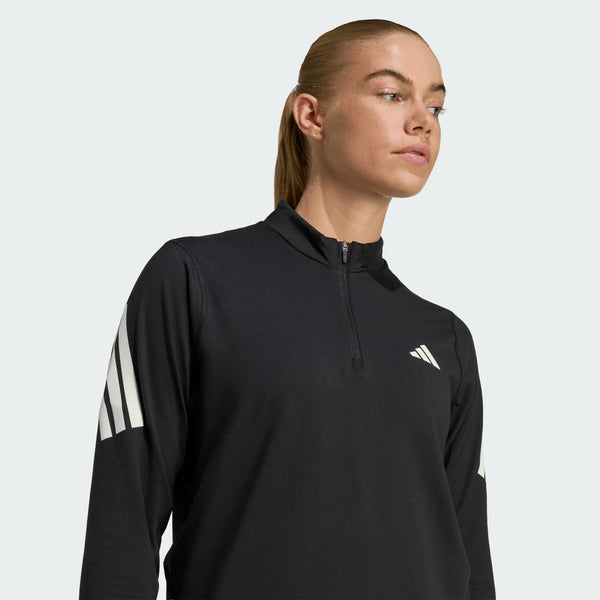 En kvinde står model for Adidas Adi365 Zip Long Sleeve Shirt i sort med hvide ærmestriber og en kvart lynlås på en almindelig lys baggrund og kigger lidt til siden.