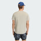 En person står med ryggen til kameraet iført en beige Adidas Adi365 Running Iconic T-Shirt (Herre, Woca), blå og sorte sportsshorts og en blå baseballkasket på en lysegrå baggrund.
