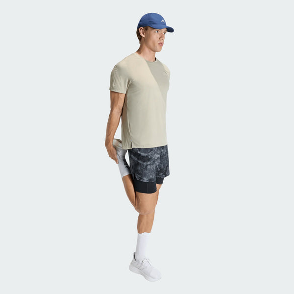 En mand iført Adidas Adi365 Running Iconic T-shirt, mønstrede shorts, hvide sokker og sneakers samt en blå kasket strækker sit ben.