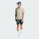 En mand står på en almindelig lys baggrund iført en Adidas Adi365 Running Iconic T-Shirt (Herre, Woca) i beige, sorte/grå mønstrede shorts over sorte kompressionsshorts, hvide strømper, hvide adidas-sneakers og en blå kasket.