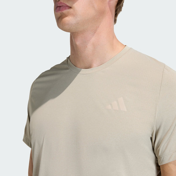 En person er iført en beige Adidas Adi365 Running Iconic T-Shirt, Herre, Woca, med et diskret logo på venstre bryst. Billedet viser overkroppen og en del af ansigtet mod en almindelig lys baggrund.
