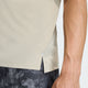 Et nærbillede viser en person iført en Adidas Adi365 Running Iconic T-Shirt, Herre, Woca i lys beige med mørke mønstrede shorts. Billedet fremhæver trøjens slids i siden nær kanten og personens arm.