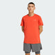 En person kigger fremad iført Adidas Adi365 Running Tee, Herre, Duor - en lys orange løbetrøje med et lille logo på brystet og grå shorts. Hænderne er bag ryggen mod en almindelig lysegrå baggrund, perfekt til løbetræning.