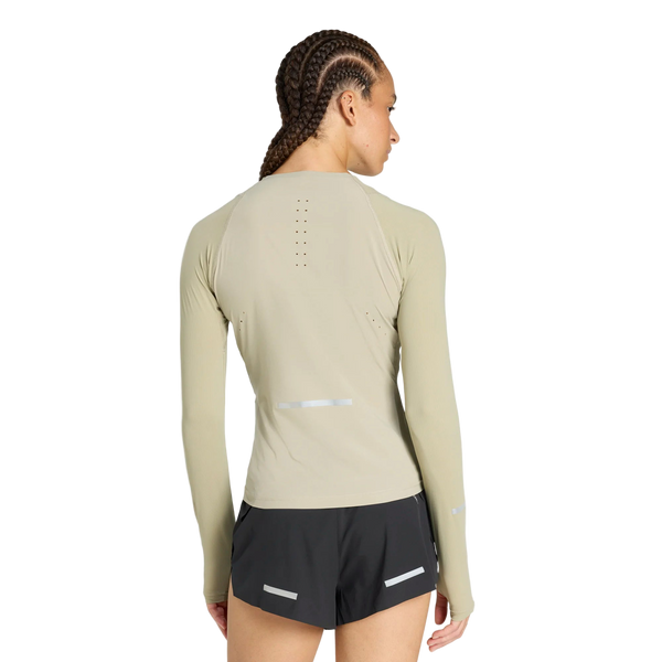 En kvinde med flettet hår ses bagfra i en tætsiddende beige Adidas Adi365 Long Sleeve Shirt, dame, woca, med små ventilationshuller, parret med sorte shorts med refleksstriber på en almindelig hvid baggrund.