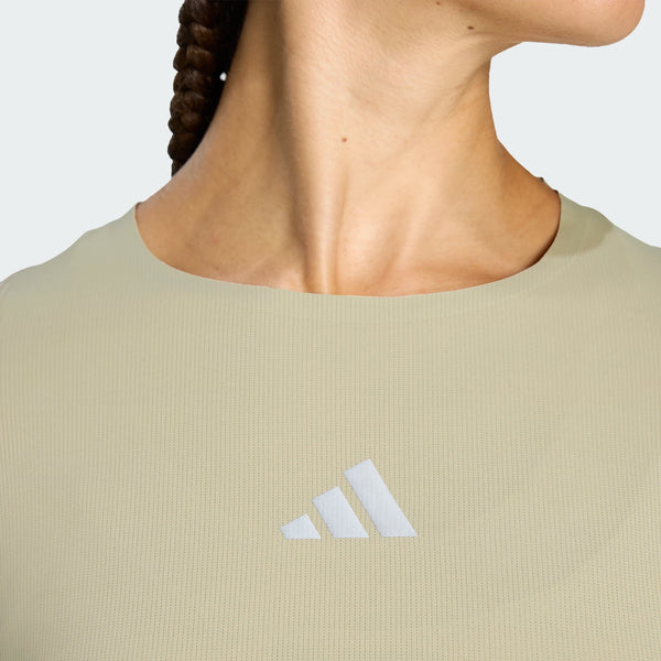 Et nærbillede viser en person iført Adidas Adi365 Long Sleeve Shirt, Dame, Woca i lys beige med det hvide Adidas-logo centreret på brystet. Personens hoved er let drejet, og det flettede hår er delvist synligt.