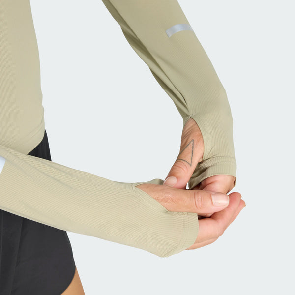 En kvinde iført en beige Adidas Adi365 Long Sleeve Shirt, Dame, Woca med tommelfingerhuller og sorte shorts justerer ærmet. Trøjen har struktureret stof og reflekterende ærmedetaljer.