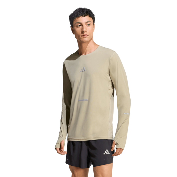 En mand er iført en lys beige Adidas Adi365 Long Sleeve Shirt, Herre, Woca med huller til tommelfingrene og sorte sportsshorts, begge med små Adidas-logoer, på en almindelig hvid baggrund.