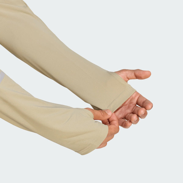 En person justerer manchetten på en beige Adidas Adi365 Long Sleeve Shirt, Herre, Woca med huller til tommelfingrene og viser begge hænder mod en almindelig lys baggrund.