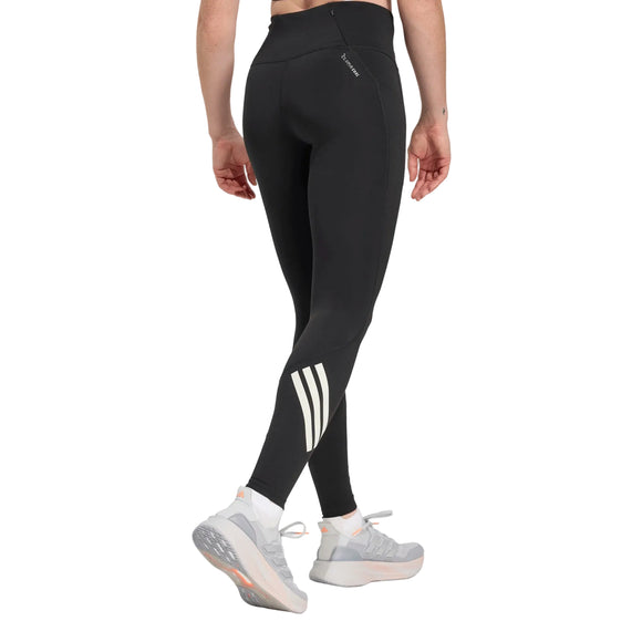 En kvinde ses fra taljen og ned iført Adidas Adi365 3S 1/1 Leggings i sort med tre hvide striber på det nederste venstre ben, sammen med lysegrå sportssko med hvide såler på en almindelig lys baggrund.