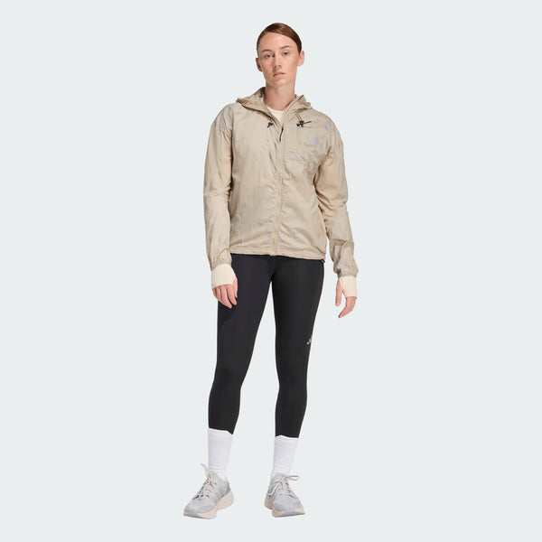 En kvinde ser fremad i Adi365 3S 1/1 Leggings til kvinder fra Adidas i sort, stylet med en lys beige jakke med lynlås, hvide strømper og grå sportssko på en almindelig lys baggrund.