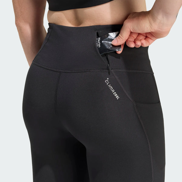 En kvinde iført Adidas Adi365 3S 1/1 Leggings i sort stikker en lille sort genstand mærket Adidas ned i den diskrete baglomme i linningen. Leggingsene har synlige sømme og "Climacool" trykt på siden.