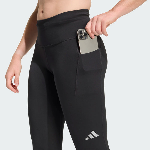 En kvinde iført Adidas Adi365 3S 1/1 Leggings i sort lægger en smartphone i sidelommen; leggingsene har et lille hvidt Adidas-logo på låret.