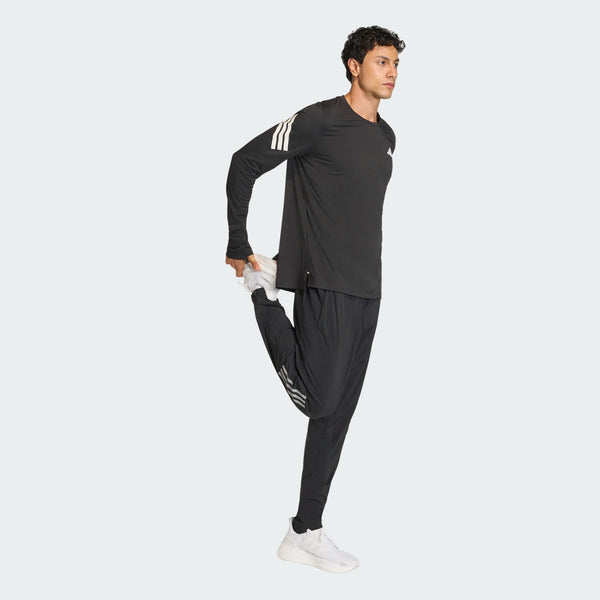 En mand iført Adidas Adi365 Long Sleeve Shirt i sort balancerer på det ene ben med den anden fod trukket tilbage i en quad stretch mod en almindelig lys baggrund.